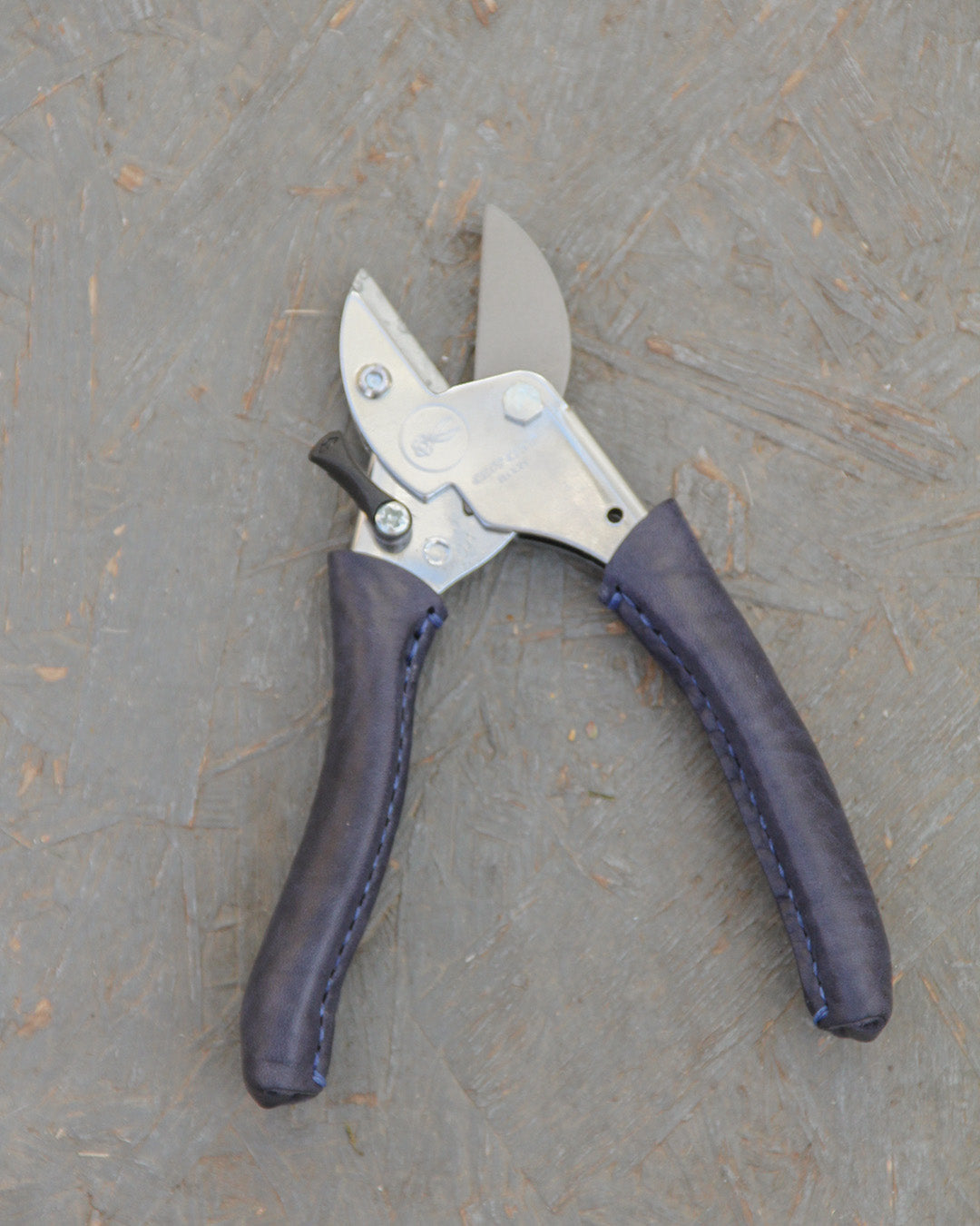 Die Chop Chop Bloom Chop Chop | Exklusive Gartenschere 'Poppy Blue' mit Metallklingen und dunkelblauen, handgenähten Griffen ruht auf einer strukturierten, gräulichen Oberfläche.