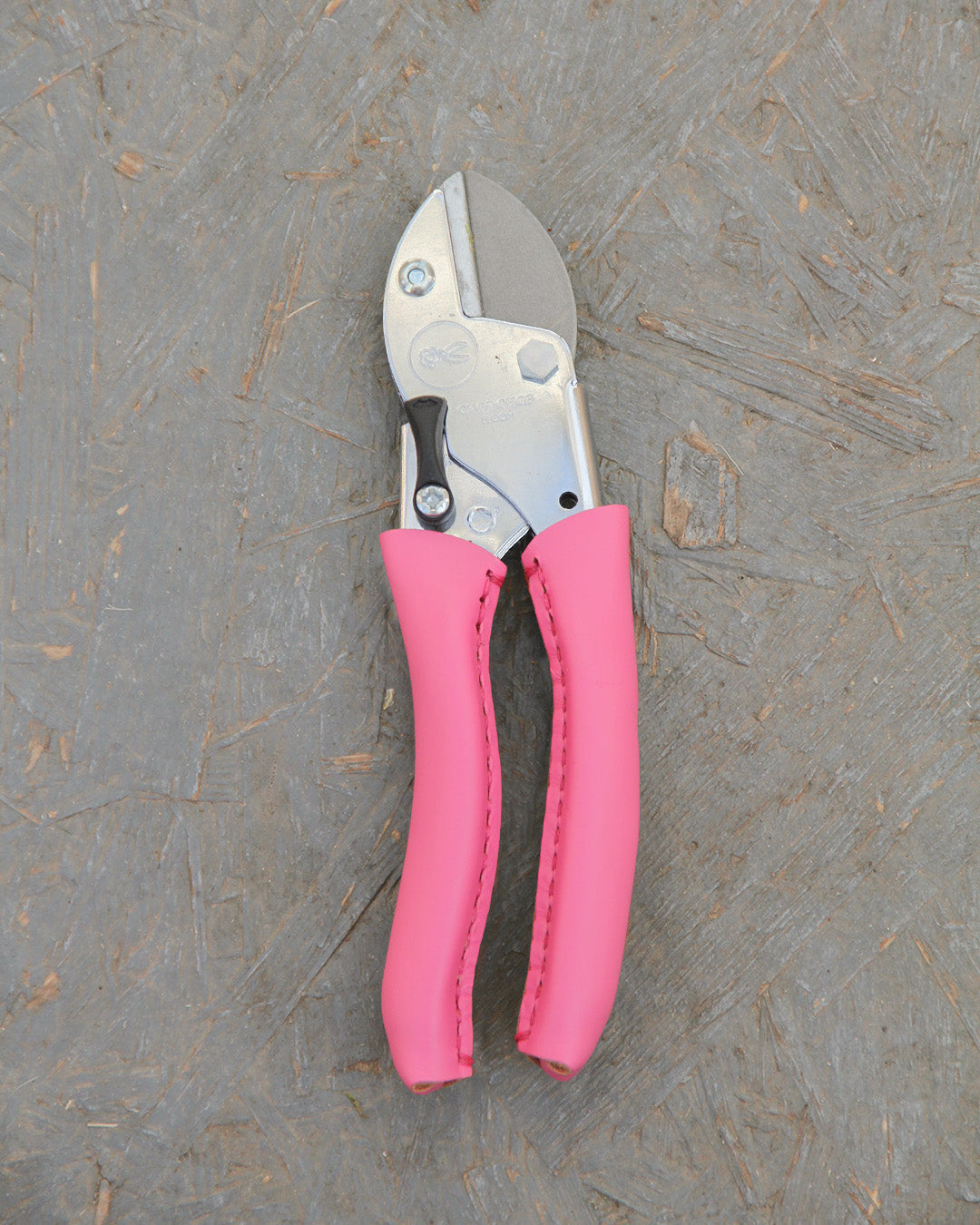 Die Chop Chop Bloom Hochwertige Gartenschere 'Pink Tulip' verfügt über Metallklingen und leuchtend pinke Gummigriffe, inspiriert vom Symbol der Initiative Brustkrebs, und ruht auf einer strukturierten grauen Holzoberfläche.