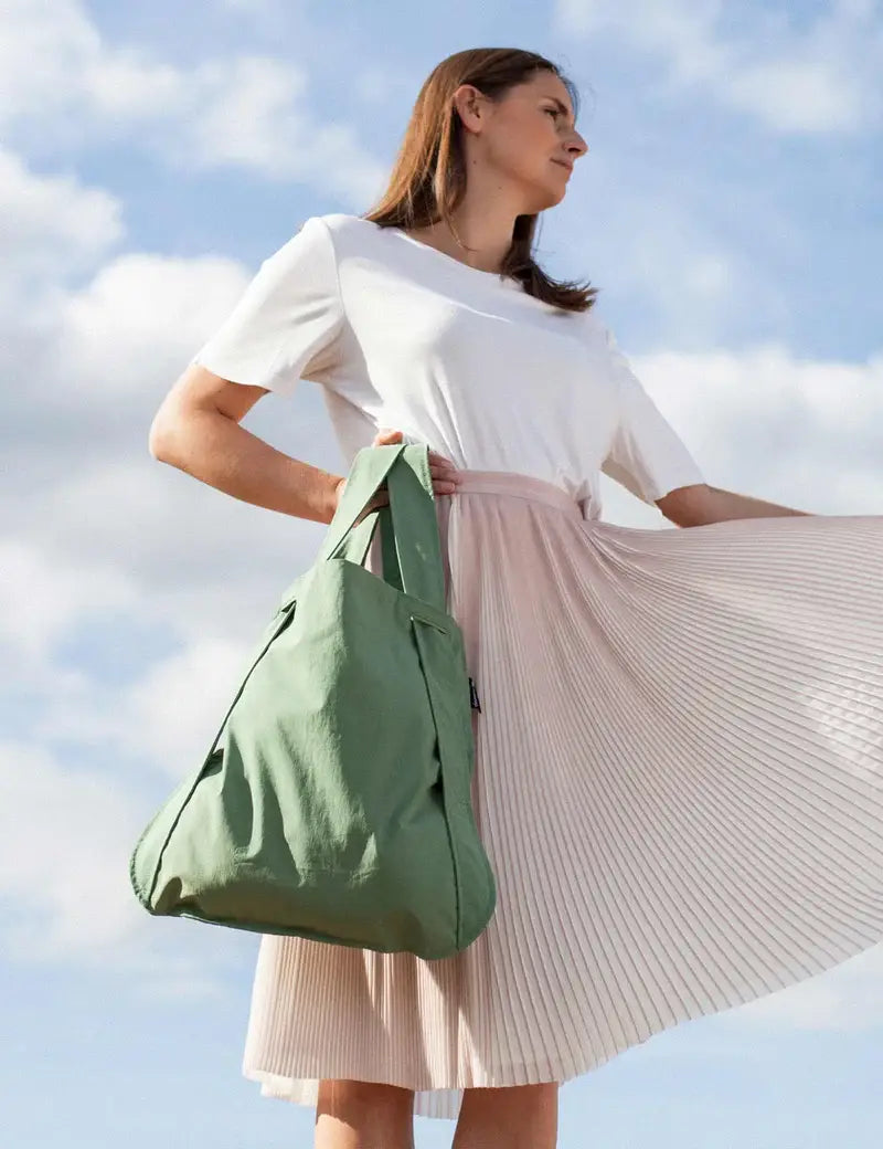 Eine Frau in einem weißen Oberteil und einem hellrosa Faltenrock hält mit einem Arm eine Notabag | Rucksack und Tasche in einem „Olive“ von Notabag in der Hand, während sie draußen vor einem blauen Himmel mit Wolken steht. Diese stilvolle, wiederverwendbare Tasche vereint mühelos Funktionalität und Eleganz für jeden Anlass.