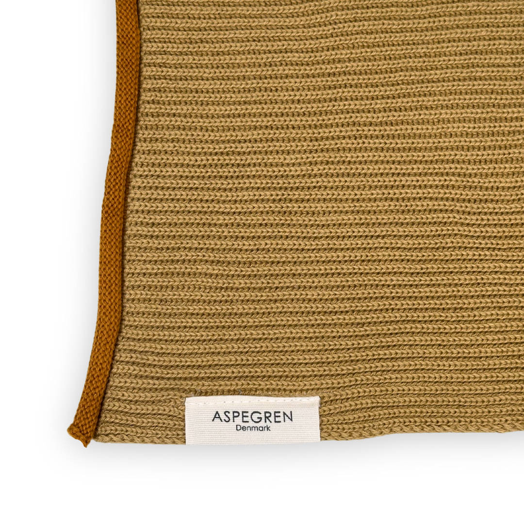 Eine Nahaufnahme eines beigen Strickstoffs mit braunem Rand, auf dessen Unterseite ein weißes Etikett mit der Aufschrift „Aspegren Denmark“ zu sehen ist. Dieses Handtuch aus Bio-Baumwolle ist Teil des „Aspegren Denmark | Küchentuch 3er-Geschenksets in Naturtönen“ – eine ideale, nachhaltige Ergänzung für Ihre Küchentextilien-Kollektion.
