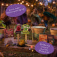 Eine Halloween-Szene im Freien mit kostümierten Kindern, die im Hintergrund tanzen, und einer farbenfrohen Auslage festlicher Snacks und Getränke mit den Bezeichnungen „Monster-Limo“, „Monsterlolly“ und „Hexeneier“ – ergänzt durch die kreative Einbindung von Stadtgärtners Saatgut „Süßes oder Saures“ | Halloween-Saatpille „Süßes“ sowie eine dekorative Anordnung aus „Wilde Saatgutmischung“ für einen zusätzlichen Hauch von Herbstzauber.
