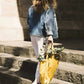 Eine Person mit welligem braunem Haar, die eine Jeansjacke, eine weiße Hose und Turnschuhe trägt, steigt eine Steintreppe hinauf und trägt dabei die Notabag „Original Golden“ – eine vielseitige Tasche und einen Rucksack von Notabag –, die mit Eukalyptuszweigen gefüllt ist.