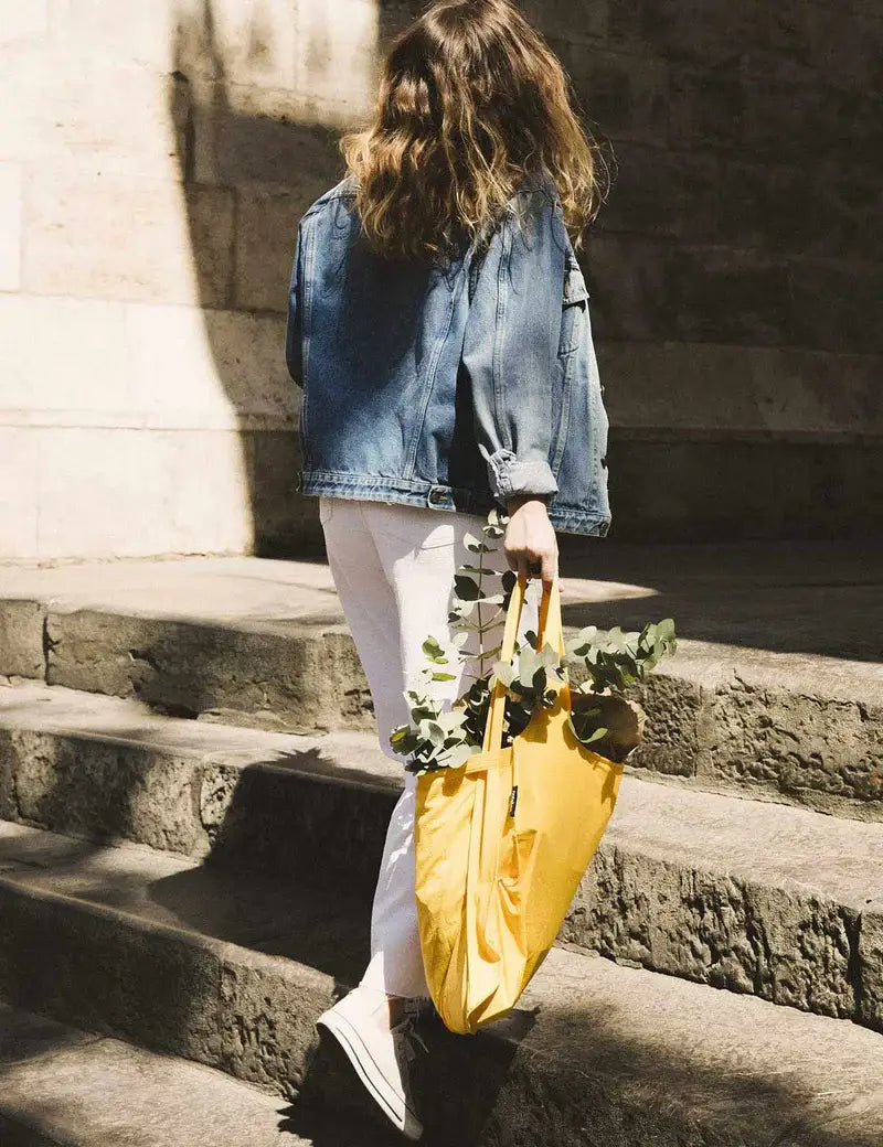 Eine Person mit welligem braunem Haar, die eine Jeansjacke, eine weiße Hose und Turnschuhe trägt, steigt eine Steintreppe hinauf und trägt dabei die Notabag „Original Golden“ – eine vielseitige Tasche und einen Rucksack von Notabag –, die mit Eukalyptuszweigen gefüllt ist.