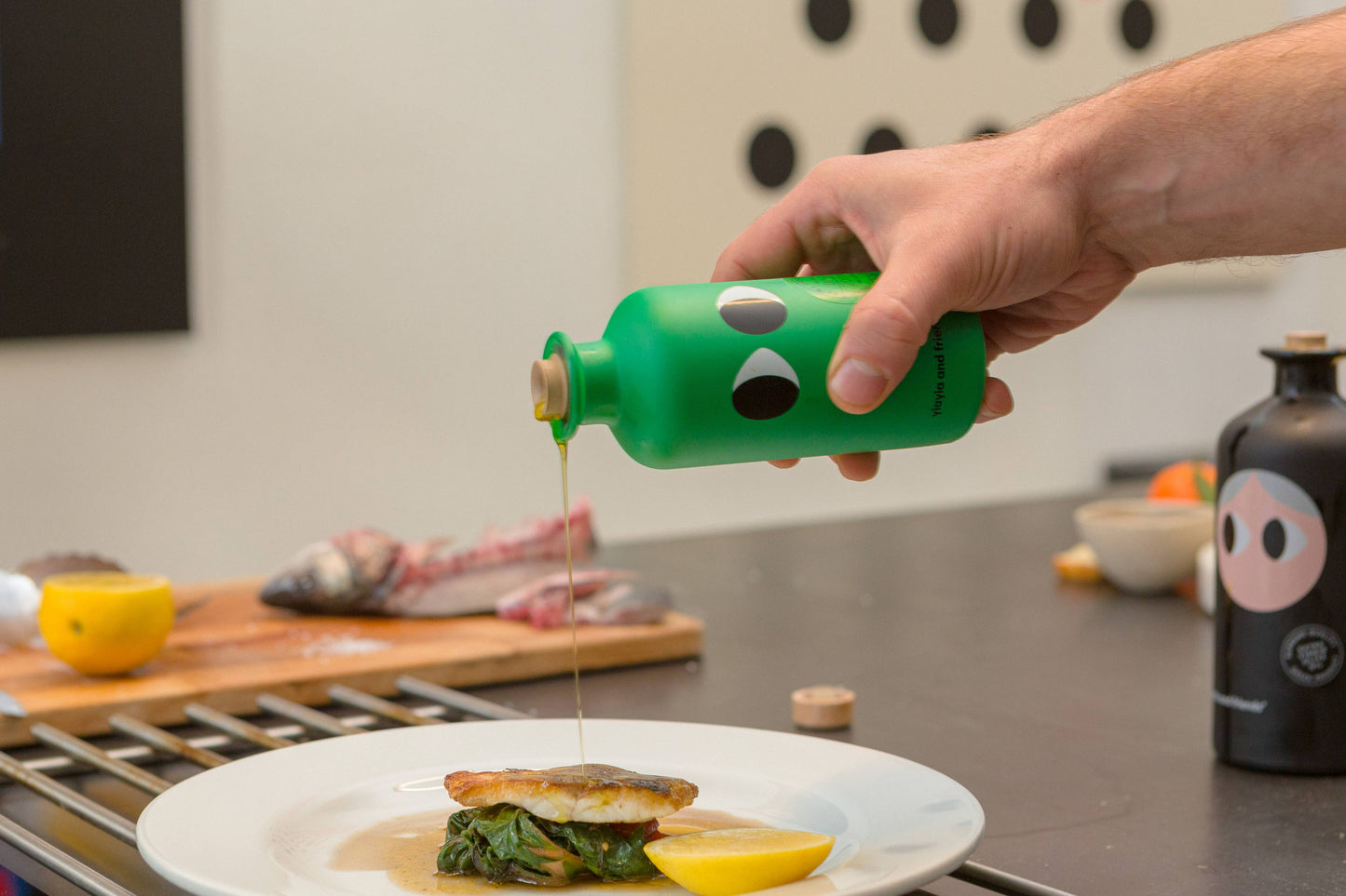 Eine Hand gießt Yiayia and friends | Natives Olivenöl extra mit Basilikum 200 ml über Fisch und Spinat auf einen Teller mit einer Zitronenspalte. Im Hintergrund stehen frischer Fisch, Zitrone und Feinkost-Zutaten von yiayia and friends auf dem Tresen.