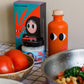 Eine leuchtende Flasche yiayia and friends | Natives Olivenöl extra mit Chili und ein schrulliges Glas stehen neben einer Schachtel mit abstrakten Schriftzeichen, umgeben von frischen Tomaten, einer Schüssel und einer Pfanne mit Kichererbsen und Gemüse auf einem karierten Tuch.