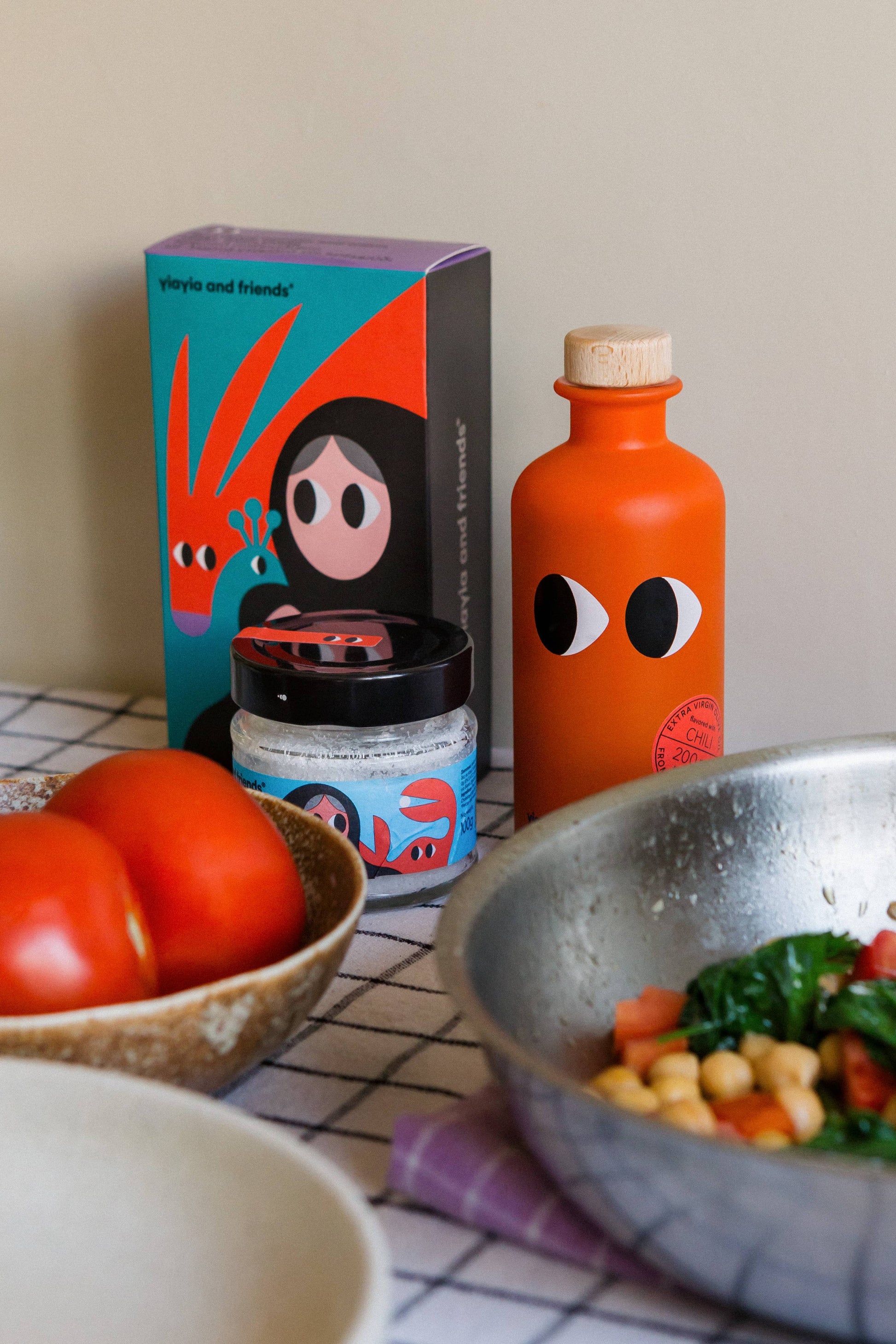 Eine leuchtende Flasche yiayia and friends | Natives Olivenöl extra mit Chili und ein schrulliges Glas stehen neben einer Schachtel mit abstrakten Schriftzeichen, umgeben von frischen Tomaten, einer Schüssel und einer Pfanne mit Kichererbsen und Gemüse auf einem karierten Tuch.