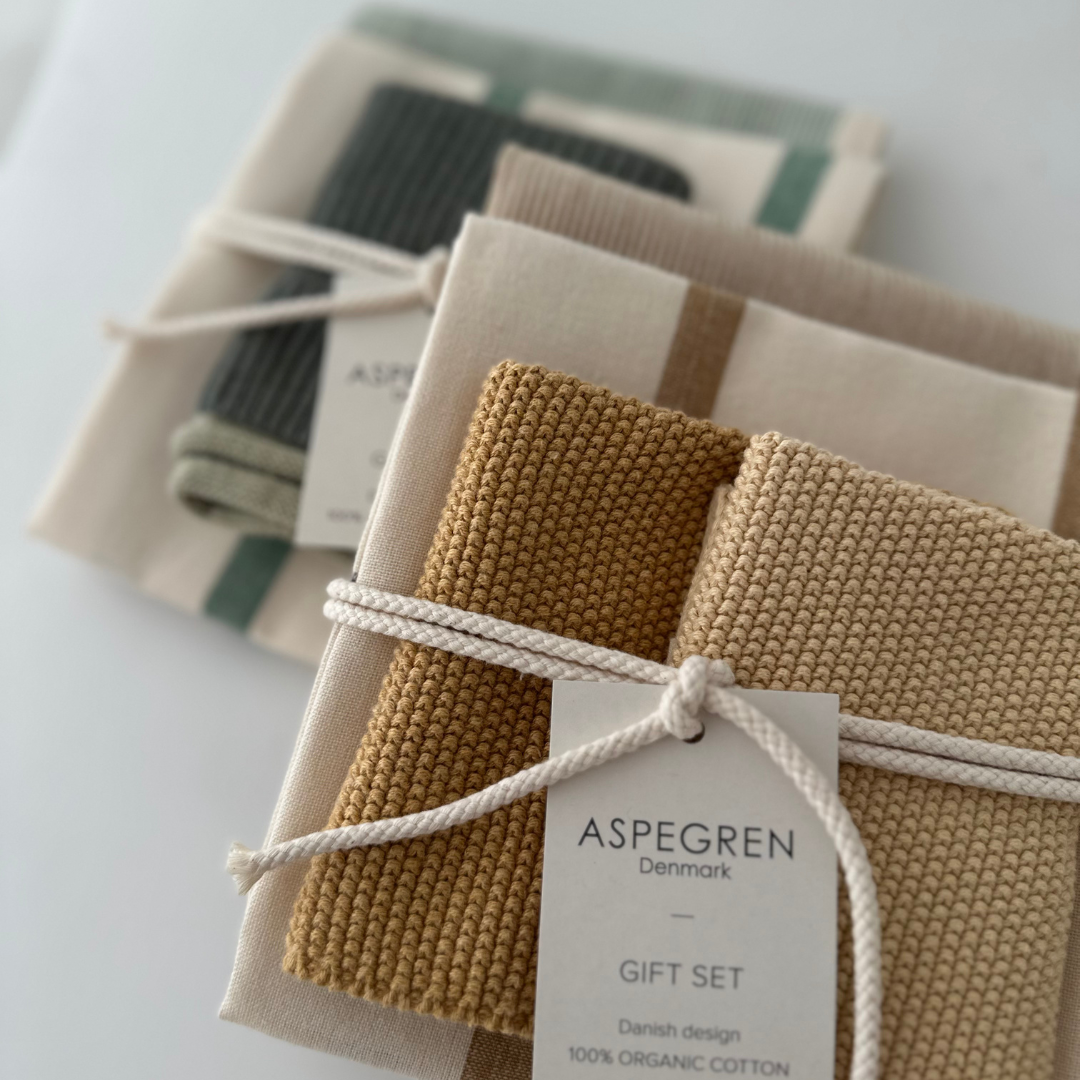 Zwei Sets des „Grünes Küchentuch 3er Geschenkset“ von Aspegren Denmark, die jeweils aus gefalteten, in neutralen Farbtönen gehaltenen Küchentüchern aus Bio-Baumwolle bestehen, mit einer weißen Kordel zusammengebunden und mit einem Geschenkanhänger von Aspegren Denmark versehen sind, sind ordentlich auf einer weißen Fläche angeordnet.