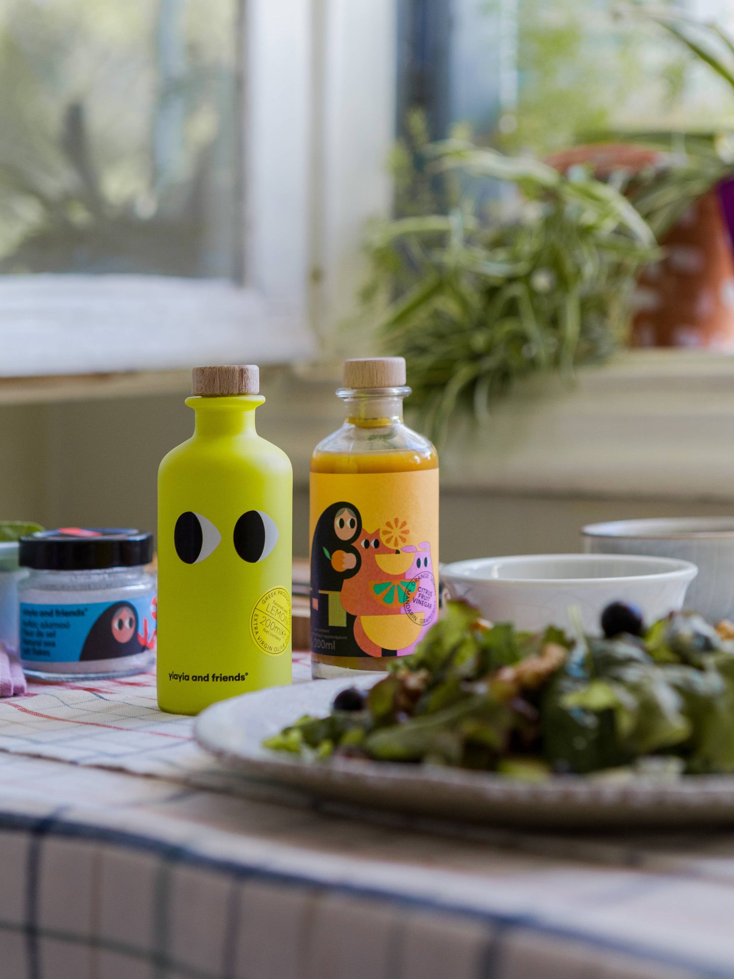 Zwei leuchtende Flaschen, darunter Yiayia and friends Natives Olivenöl extra mit Zitrone 200 ml, stehen auf einem Tisch mit einem Glas, einer Schale und einem Salatteller. Sonnenlicht und Pflanzen im Hintergrund sorgen für eine helle, lässige Feinkost-Stimmung.