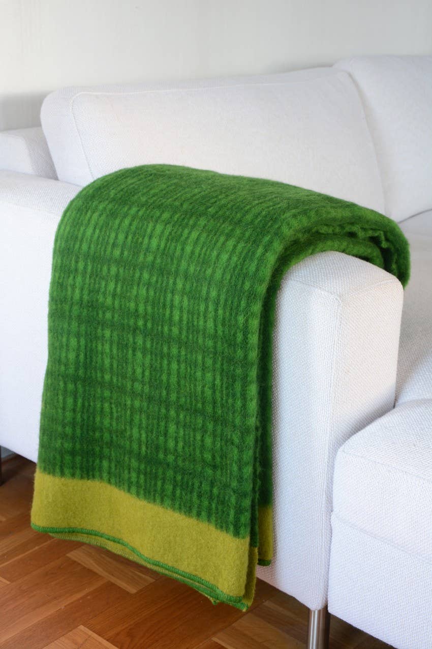 Eine Sofie Sjöström Design 'Darling' grüne Tagesdecke aus Lammwolle mit gelbem Rand liegt über dem Arm eines weißen Sofas auf einem Holzboden.
