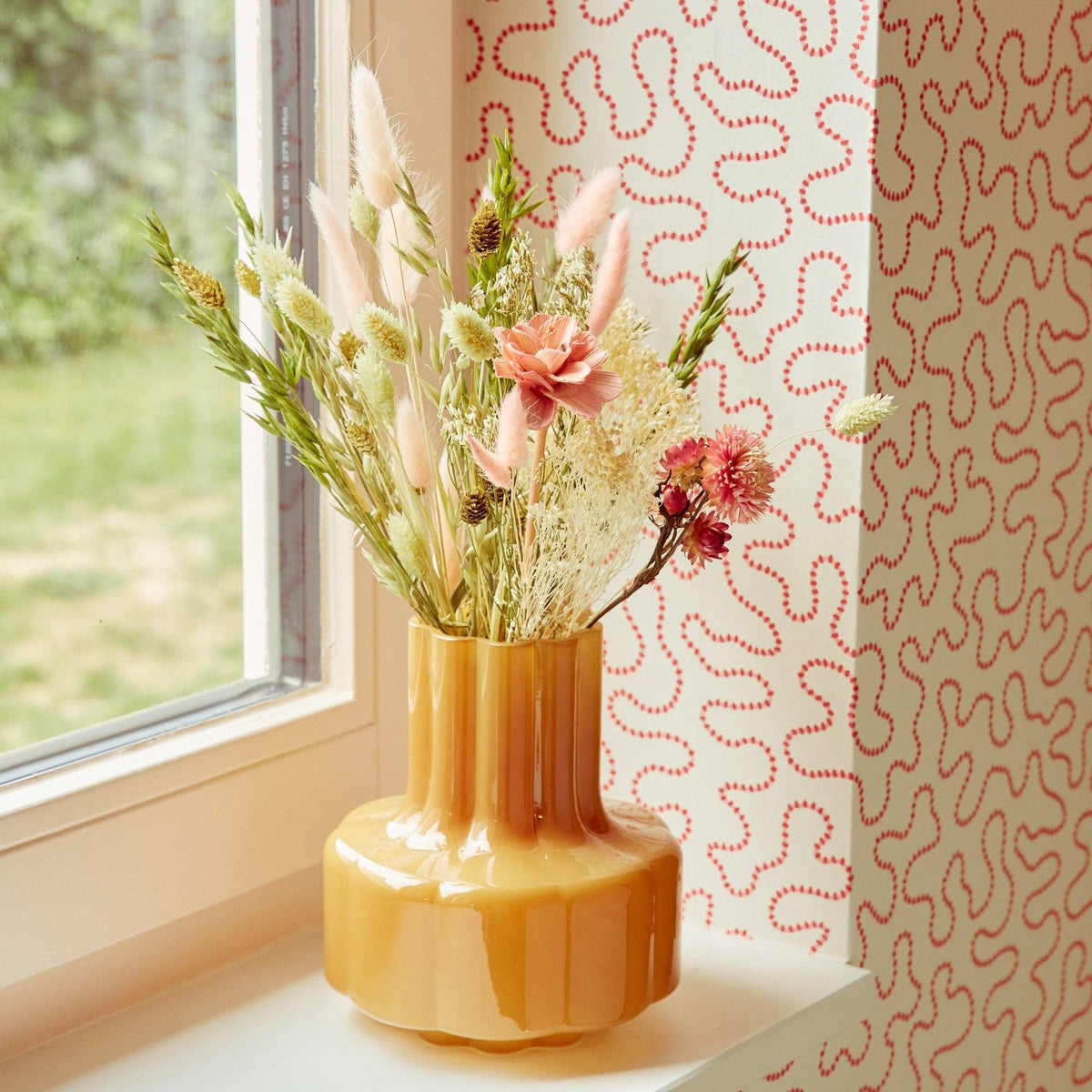 Auf einer Fensterbank steht eine gelbe Vase mit dem Feldstrauß „Rosa Eleganz“ (Größe S) von Wildflowers by Floriette. Die Wand dahinter ist mit einer Tapete mit verschnörkelten Mustern verziert, und Sonnenlicht taucht die Szene in helles Licht.