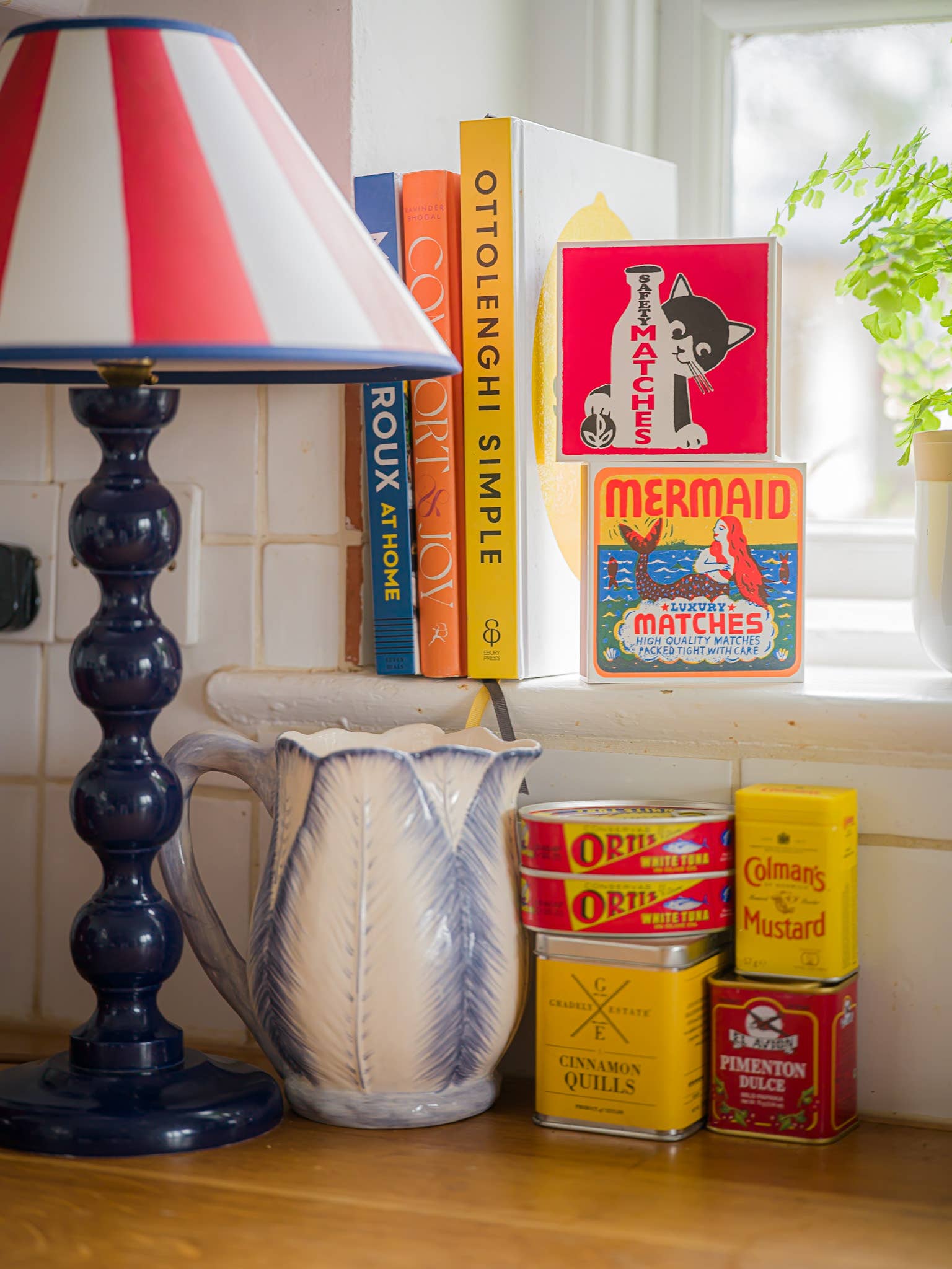 In einer gemütlichen Küchenecke stehen eine blau-weiße Lampe, Kochbücher, die Streichholzschachtel "Mermaid" Quadrat Streichhölzer von Archivist Gallery, Gewürzdosen, ein Keramikkrug und eine Topfpflanze auf der Fensterbank. Sonnenlicht durchflutet den einladenden Raum.