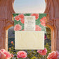 Ein Barren Panier Des Sens Duftseife Rose des Bastides 150 g steht vor seinem Blumenkasten, umrahmt von einem steinernen Torbogen mit Blick auf rosa Rosen und Berge im Sonnenuntergang.