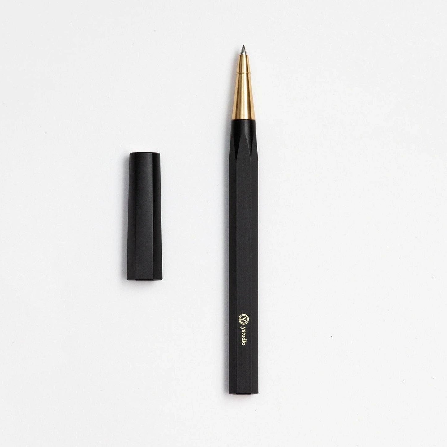 Der ENTREPÔT YSTUDIO | Harz-Rollerball-Stift (Schwarz) mit goldener Spitze und schwarzer Kappe präsentiert sich im schlichten Acryl- und Messing-Design mit Logo und Text in Bodennähe auf weißem Untergrund.