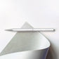 Der ENTREPÔT YSTUDIO Klassischer Revolve Kugelschreiber Slim (Glänzendes Silber) liegt waagerecht auf einer weißen Fläche neben einem teils eingerollten Blatt mit sanft grauer Unterseite.