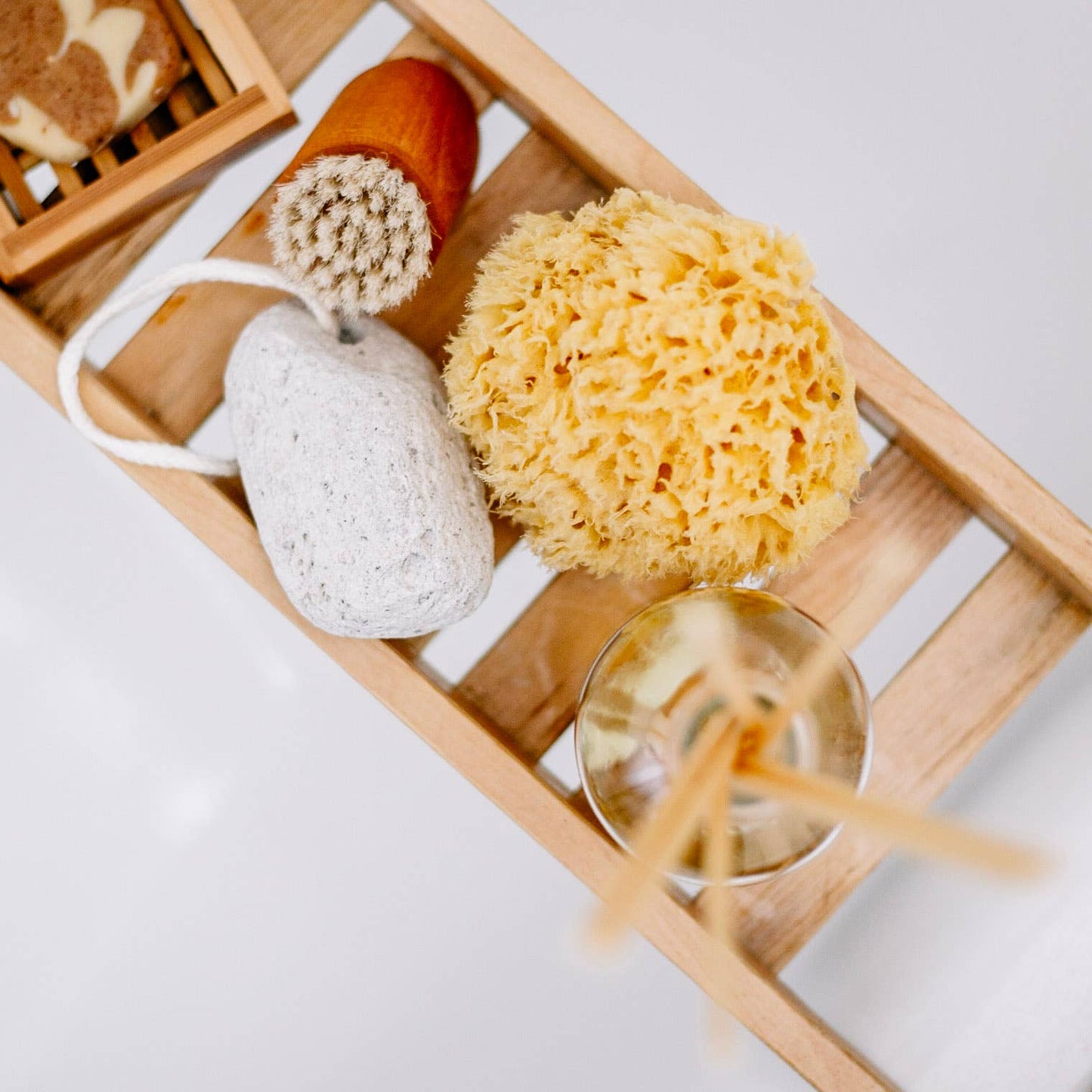 Eine hölzerne Badeschale enthält einen Croll & Denecke nachhaltigen Naturschwamm aus dem Meer S, einen Bimsstein, eine Bürste mit Holzgriff, Seife in einer Schale und ein Glas mit Schilfrohr-Diffusorstäbchen für nachhaltige Pflege.