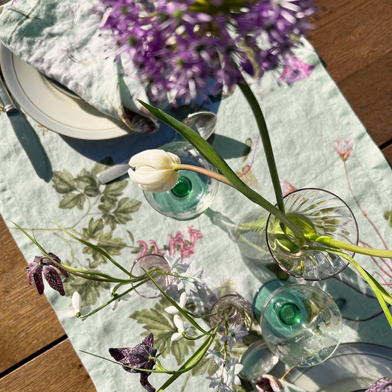 Ein sonnenbeschienener Tisch mit einem Leinen-Tischläufer „Flora Danica Atlas“ von Koustrupco (145 x 45 cm), weißen Tellern, silbernem Besteck, grünen Gläsern und Vasen mit violetten und weißen Blumen – eine wunderschöne, von Flora Danica inspirierte Tischdekoration, von oben betrachtet.