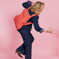 Eine Frau in dunkler Jeans und High Heels nimmt vor einem rosa Hintergrund eine verspielte Pose auf einem Bein ein und trägt dabei die „Notabag Wandelbare Tasche und Rucksack ‚Beeren‘“ von Notabag – eine innovative Tasche, die sich nahtlos von einer Einkaufstasche in einen Rucksack verwandeln lässt – über der Schulter.