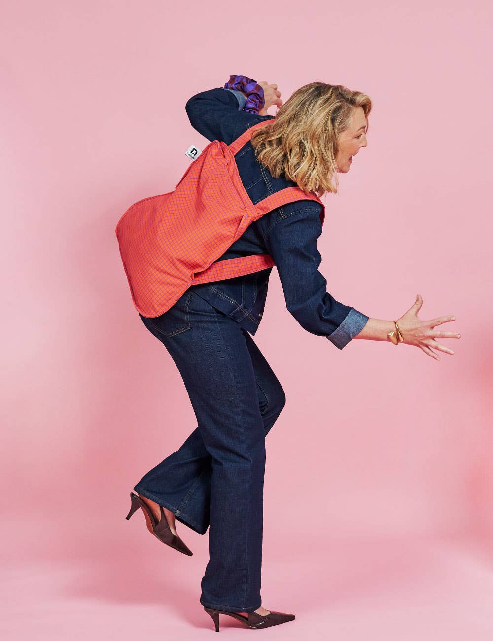 Eine Frau in dunkler Jeans und High Heels nimmt vor einem rosa Hintergrund eine verspielte Pose auf einem Bein ein und trägt dabei die „Notabag Wandelbare Tasche und Rucksack ‚Beeren‘“ von Notabag – eine innovative Tasche, die sich nahtlos von einer Einkaufstasche in einen Rucksack verwandeln lässt – über der Schulter.