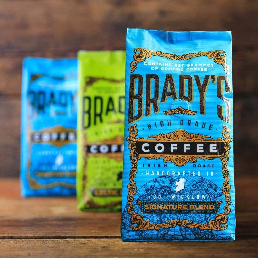 Drei stehende Tüten Brady's Coffee Company | 227 g gemahlener Signature Blend Kaffee sind auf einer Holzfläche aufgereiht. Die vordere blaue Tüte ist im Fokus, während zwei andere im Hintergrund unscharf erscheinen.