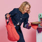 Eine lächelnde Person in Jeanskleidung hält in der einen Hand eine rote Notabag｜Wandelbare Tasche und Rucksack „Beeren“ – ein cleveres Notabag-Design, das sich von einer Einkaufstasche in einen Rucksack verwandeln lässt – und in der anderen Hand eine Torte auf einem Tuch; sie steht neben einem mintgrünen Haushaltsgerät vor einem rosa Hintergrund.