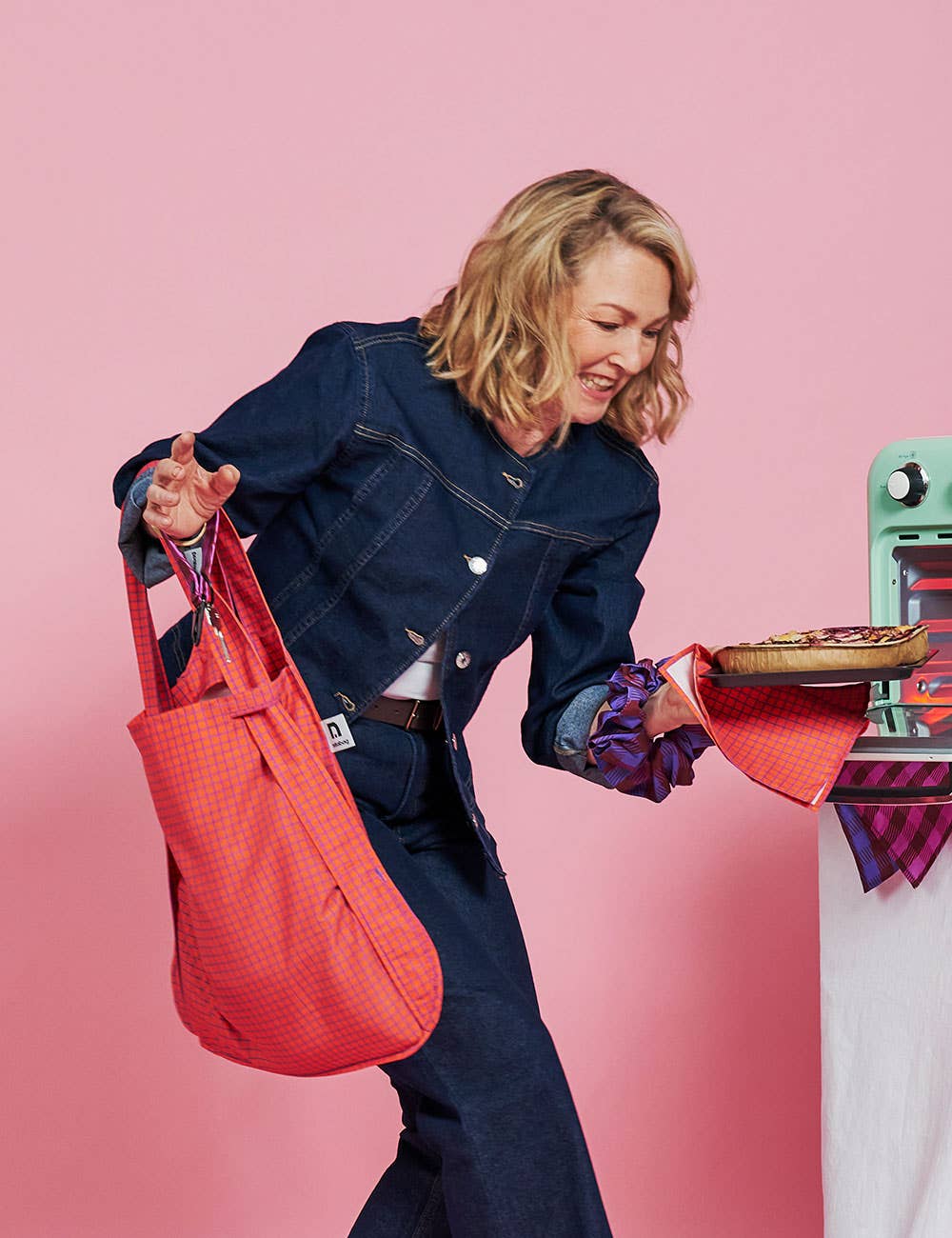 Eine lächelnde Person in Jeanskleidung hält in der einen Hand eine rote Notabag｜Wandelbare Tasche und Rucksack „Beeren“ – ein cleveres Notabag-Design, das sich von einer Einkaufstasche in einen Rucksack verwandeln lässt – und in der anderen Hand eine Torte auf einem Tuch; sie steht neben einem mintgrünen Haushaltsgerät vor einem rosa Hintergrund.