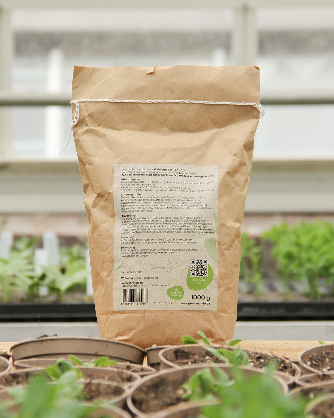 Ein 1000g-Sack Green Mundo 'Dübli' Düngedübel liegt auf einer Holzfläche zwischen kleinen Töpfen mit Erde und Setzlingen. Im Hintergrund sind üppige grüne Pflanzen und ein verschwommenes Fenster zu sehen.