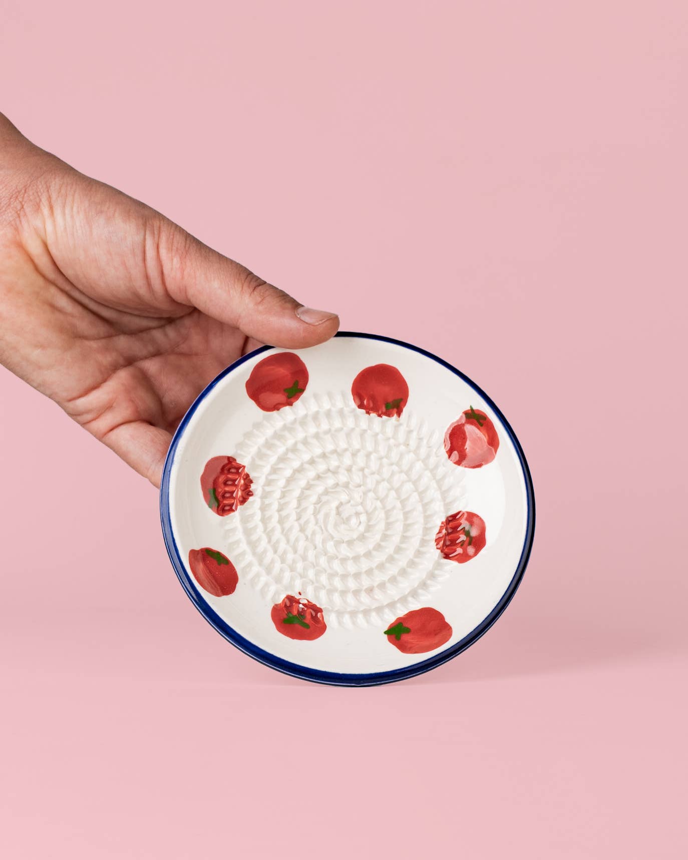 Eine Hand hält die Alfar Tierra Cocida | Kleine Keramik-Knoblauchreibe „Pomodoro“ – eine kleine, runde Knoblauchreibe aus Keramik mit einer strukturierten Spirale in der Mitte und roten Tomatenmotiven am Rand, traditionell in Spanien handgefertigt – vor einem rosa Hintergrund.