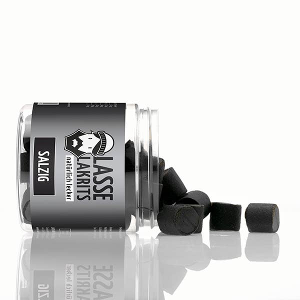 Ein durchsichtiges Glas mit der Aufschrift LASSE LAKRITS | Dose Lakritz Salzig natürlich lecker liegt auf der Seite und verschüttet mehrere Stücke des schwarzen, zylindrischen, natürlich leckeren, salzigen Lakritzbonbons der Marke auf eine weiße Fläche.