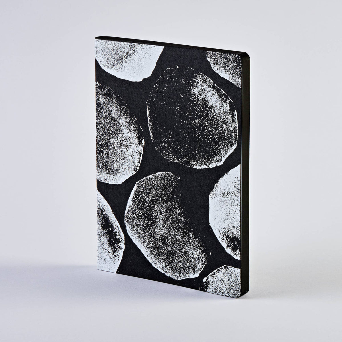Ein nuuna | „Genesis“-Notizbuch „Not Ordinary L Light“ im Format A5+ von nuuna mit schwarzem Einband und großen, weißen, strukturierten Kreismustern, das sich deutlich von einem schlichten hellgrauen Hintergrund abhebt.