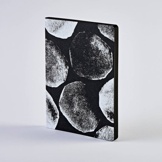 Ein nuuna | „Genesis“-Notizbuch „Not Ordinary L Light“ im Format A5+ von nuuna mit schwarzem Einband und großen, weißen, strukturierten Kreismustern, das sich deutlich von einem schlichten hellgrauen Hintergrund abhebt.