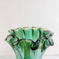 Die Murano Dream | Große „Fazzoletto“-Vase aus grünem Murano-Glas (H 32, Ø 24 cm) besticht durch ihren gewellten Rand und ihr fließendes Design. Sie steht auf hellem Untergrund vor einem schlichten Hintergrund.