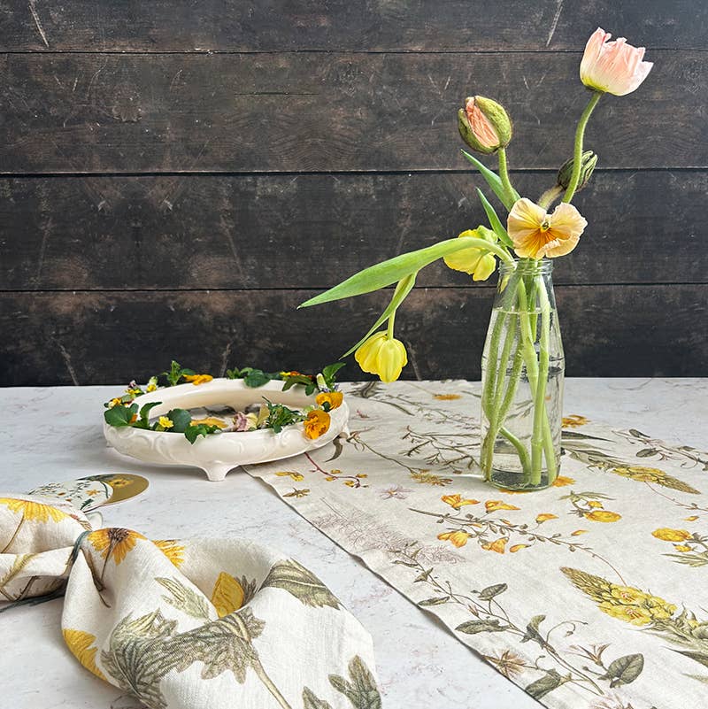 Eine Glasvase mit pastellfarbenen Blumen steht auf einem geblümten Tischläufer neben einer weißen Schale mit einem Blumenkranz und der Koustrupco-Leinenserviette mit gelbem Blütenmotiv (45 x 45 cm) und bildet so eine bezaubernde botanische Tischdekoration auf dunklem Holz.
