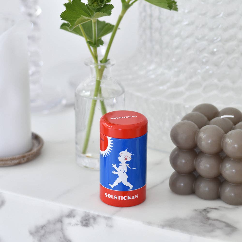 Eine blaue und rote Solstickan Design | Streichholzdose Originaldesign von Solstickan Design steht auf Marmor, begleitet von einer grauen Bubble Candle, einer weißen Kerze und einer Glasvase mit grünen Blättern.