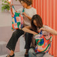 Zwei Frauen sitzen auf Betonstufen und schauen in ihre bunte Notabag | Shopping Tote 'Flow' von Notabag. Die eine hat lockiges Haar, die andere glattes. Lässig gekleidet, genießen sie ihre wiederverwendbare Tasche mit rosa Vorhängen und einer Pflanze im Rücken.