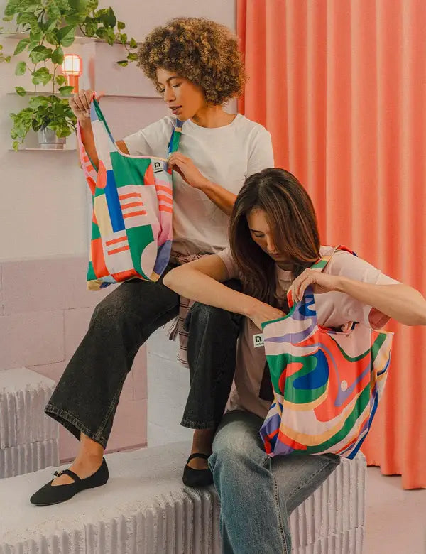 Zwei Frauen sitzen auf Betonstufen und schauen in ihre bunte Notabag | Shopping Tote 'Flow' von Notabag. Die eine hat lockiges Haar, die andere glattes. Lässig gekleidet, genießen sie ihre wiederverwendbare Tasche mit rosa Vorhängen und einer Pflanze im Rücken.