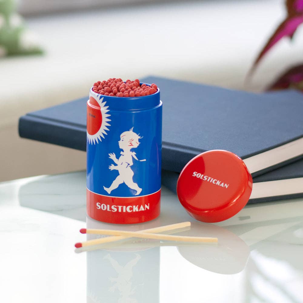 Eine Solstickan Design | Streichholzdose Originaldesign in blau und rot liegt offen, gefüllt mit extra langen Streichhölzern. Der kultige Deckel der Solstickan Design Dose liegt daneben, zusammen mit zwei Büchern und einigen losen Streichhölzern.