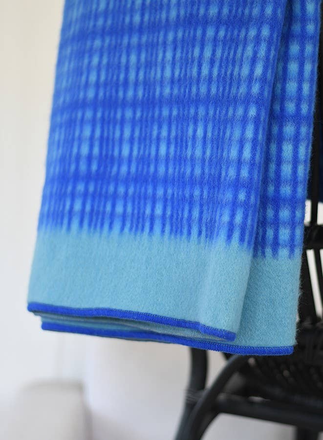 A Sofie Sjöström | 'Darling' blaue Tagesdecke aus Lammwolle von Sofie Sjöström Design mit Farbverlauf und Gittermuster liegt über einem dunklen Stuhl.