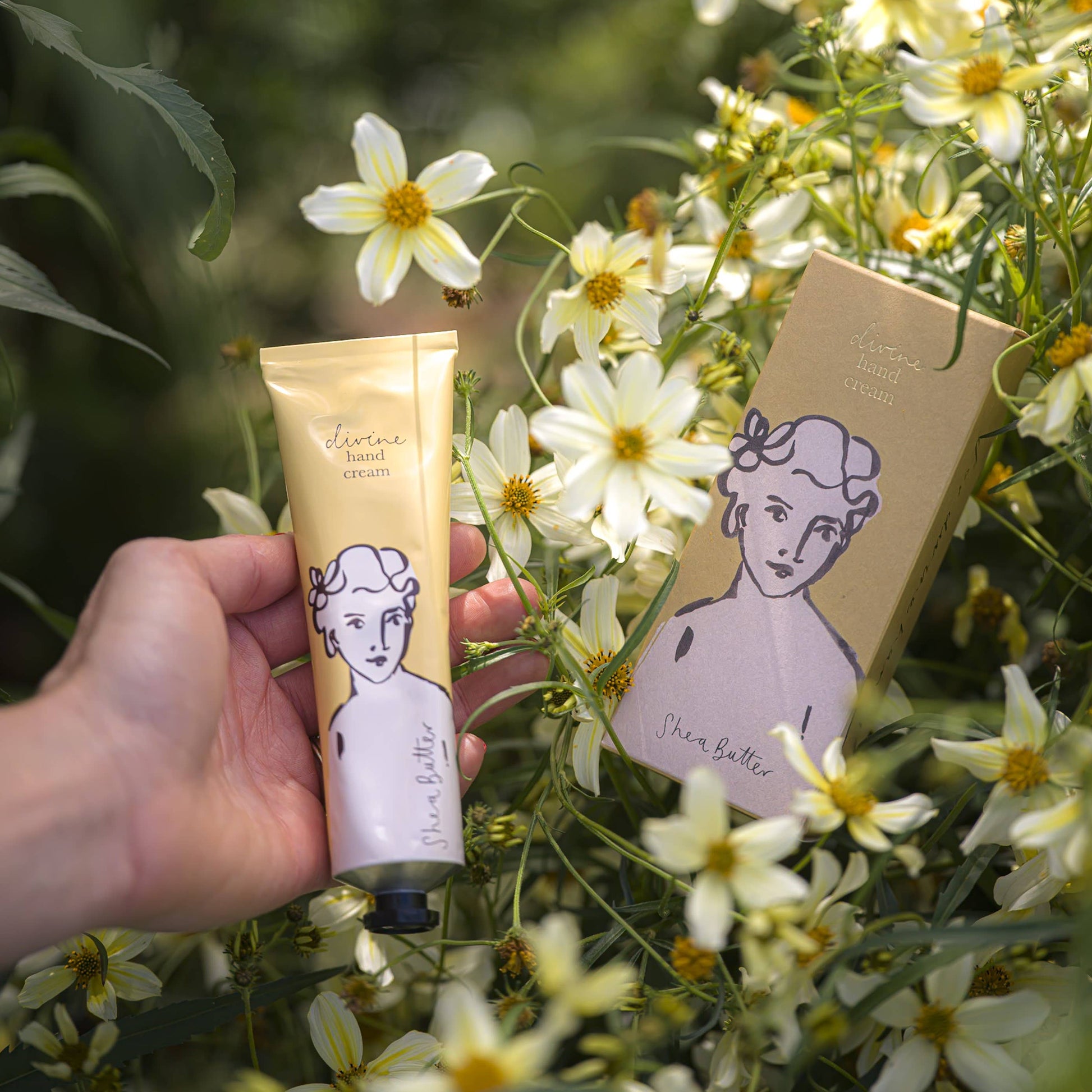 Eine Hand hält eine Tube Archivist | 'Sheabutter' Handcreme von Archivist Gallery. Auf der Verpackung ist eine Frau abgebildet, umgeben von blühenden weißen und gelben Blumen. Die passende Schachtel ruht inmitten der Blüten und unterstreicht die luxuriöse Handpflege.