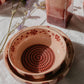 Zwei Keramikschalen von Alfar Tierra Cocida im mittelrosa „Sakura“-Design, jeweils mit handgemalten Blumen- und Spiralmotiven verziert, sind auf einer weißen, strukturierten Oberfläche gestapelt. Daneben steht eine quadratische, rot-rosa Flasche von Alfar Tierra Cocida, während hier und da Zweige mit violetten Blüten verstreut sind – perfekt, um Ihrer „Küchenhelfer“-Kollektion einen künstlerischen Touch zu verleihen.