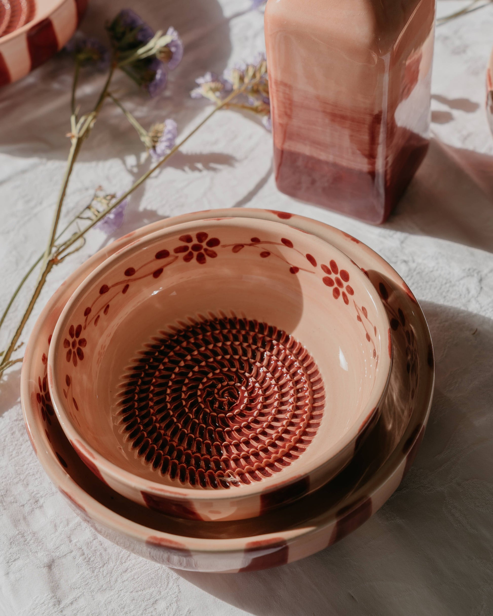 Zwei Keramikschalen von Alfar Tierra Cocida im mittelrosa „Sakura“-Design, jeweils mit handgemalten Blumen- und Spiralmotiven verziert, sind auf einer weißen, strukturierten Oberfläche gestapelt. Daneben steht eine quadratische, rot-rosa Flasche von Alfar Tierra Cocida, während hier und da Zweige mit violetten Blüten verstreut sind – perfekt, um Ihrer „Küchenhelfer“-Kollektion einen künstlerischen Touch zu verleihen.