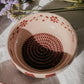 Eine mittelrosa „Sakura“-Keramikschale mit Knoblauchreibe von Alfar Tierra Cocida steht auf einer weißen Fläche in der Nähe violetter Blumen im sanften Sonnenlicht – ein handbemaltes Küchenutensil mit kunstvollem Flair, inspiriert von der spanischen Keramikkunst.