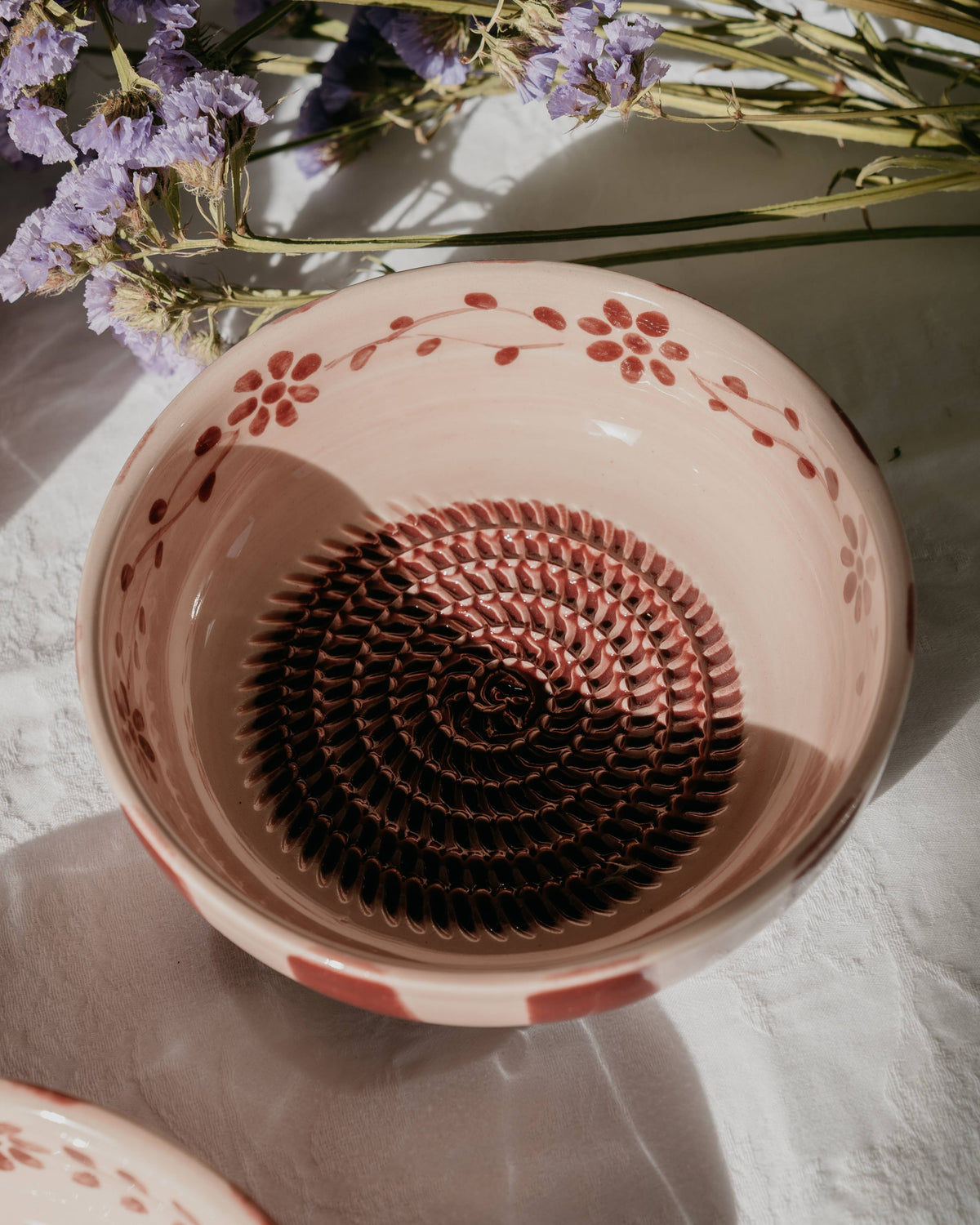 Eine mittelrosa „Sakura“-Keramikschale mit Knoblauchreibe von Alfar Tierra Cocida steht auf einer weißen Fläche in der Nähe violetter Blumen im sanften Sonnenlicht – ein handbemaltes Küchenutensil mit kunstvollem Flair, inspiriert von der spanischen Keramikkunst.