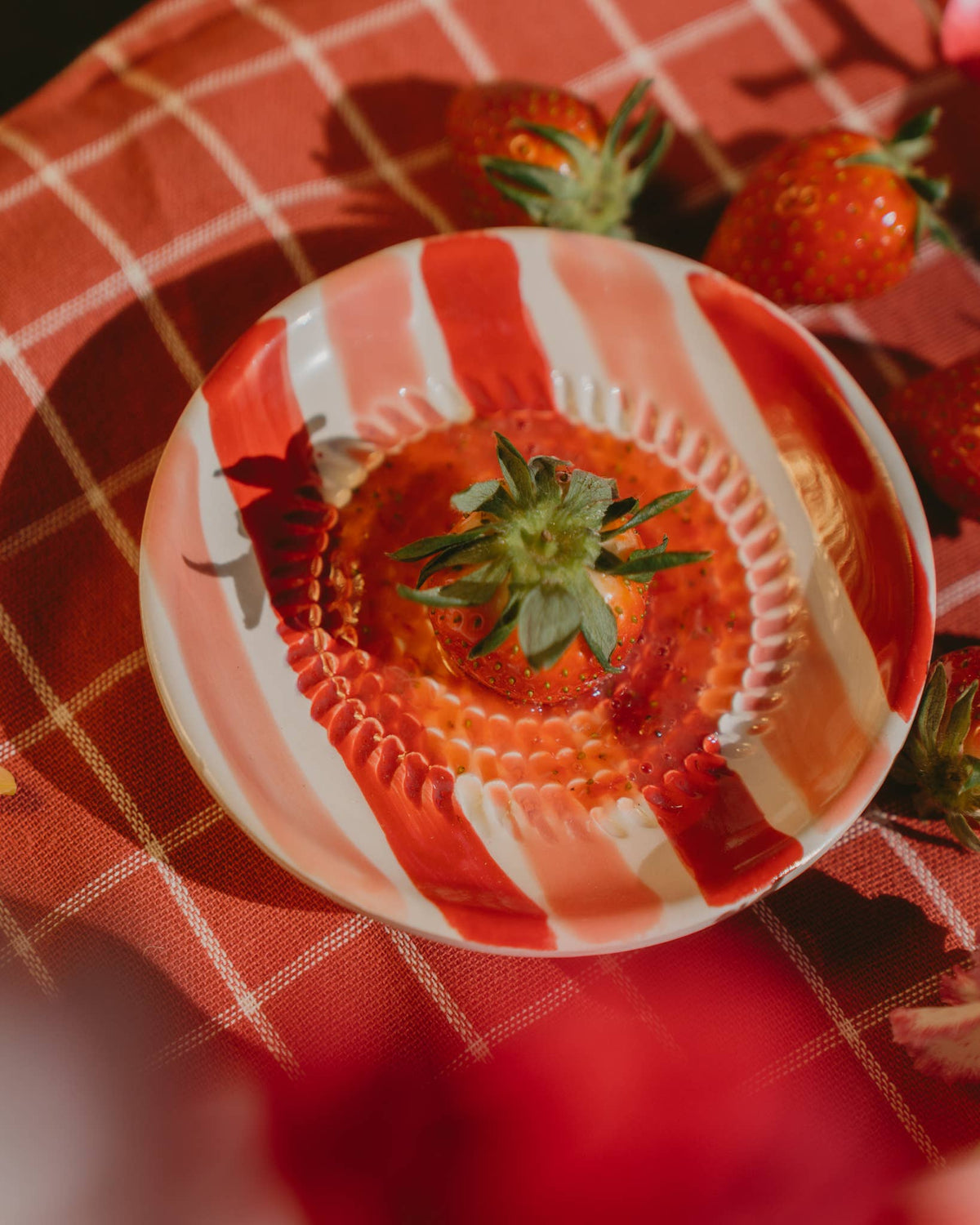 Eine ganze Erdbeere liegt in einer rot-weiß gestreiften Schale auf einer rot karierten Tischdecke, umgeben von weiteren Erdbeeren in hellem, natürlichem Licht – perfekt ergänzt durch die „Alfar Tierra Cocida | Kleine keramische Knoblauchreibe Rosa & Rot“ als charmantes Küchenaccessoire von Alfar Tierra Cocida.