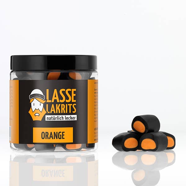 Eine LASSE LAKRITS Dose Lakritz Orange gefüllt mit fruchtiger, schwarz-oranger Lakritze ohne Palmöl sitzt neben einem kleinen Stapel der gleichen Lakritzbonbons auf einer weißen Fläche mit einfarbigem Hintergrund.