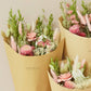 Drei in braunes Papier eingewickelte Sträuße aus rosa, grünen und weißen getrockneten Wildblumen – „Wildflowers by Floriette’s Trockenblumen ‚Rosa Eleganz‘ Feldstrauß in S“ – vor einem hellbeigen Hintergrund. Die Verpackung ist mit dem „Wildflowers by Floriette“-Logo versehen, das die Eleganz jedes einzelnen, einzigartigen Straußes unterstreicht.