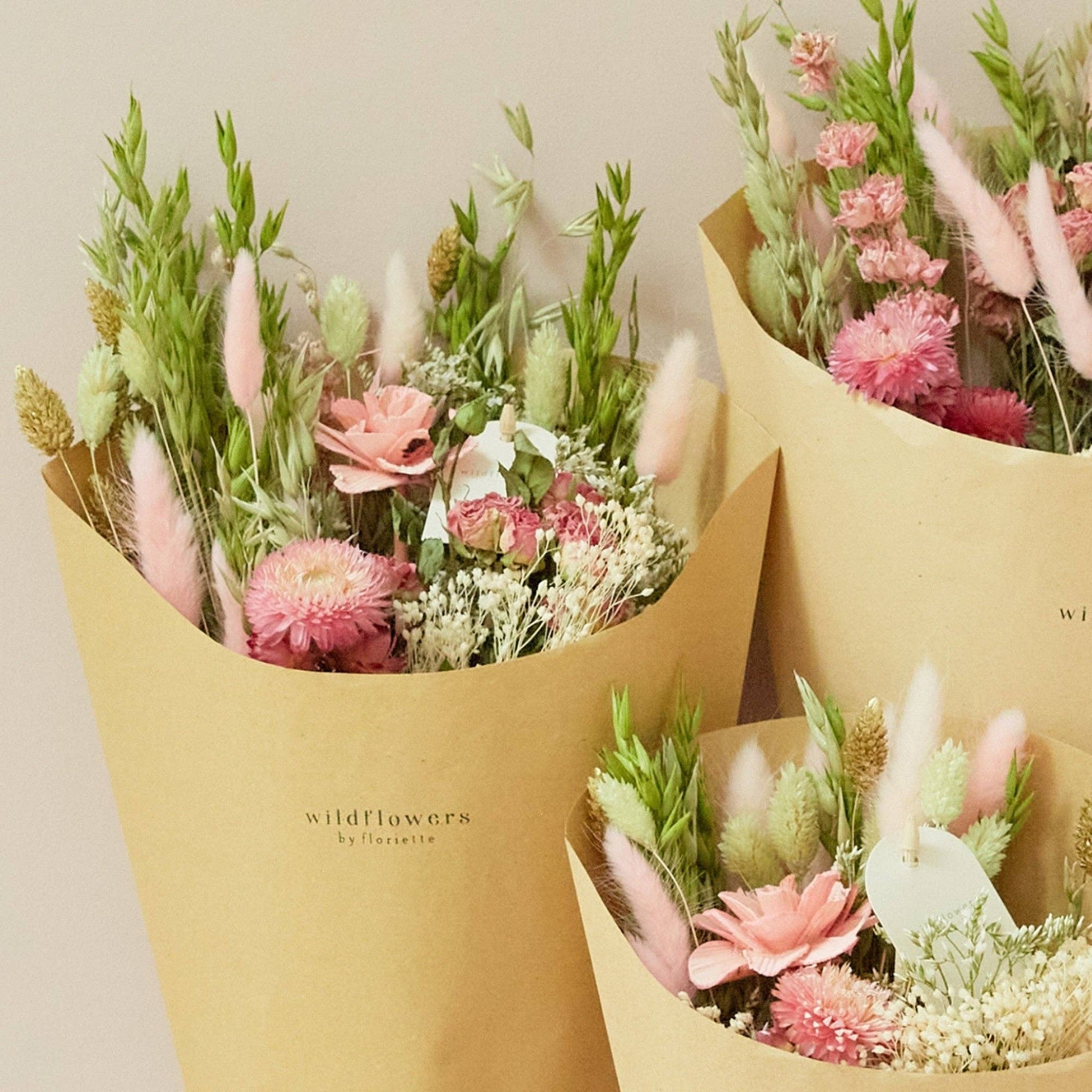 Drei in braunes Papier eingewickelte Sträuße aus rosa, grünen und weißen getrockneten Wildblumen – „Wildflowers by Floriette’s Trockenblumen ‚Rosa Eleganz‘ Feldstrauß in S“ – vor einem hellbeigen Hintergrund. Die Verpackung ist mit dem „Wildflowers by Floriette“-Logo versehen, das die Eleganz jedes einzelnen, einzigartigen Straußes unterstreicht.