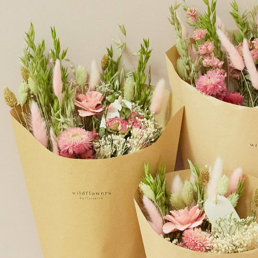 Drei in braunes Papier eingewickelte Sträuße aus rosa, grünen und weißen getrockneten Wildblumen – „Wildflowers by Floriette’s Trockenblumen ‚Rosa Eleganz‘ Feldstrauß in S“ – vor einem hellbeigen Hintergrund. Die Verpackung ist mit dem „Wildflowers by Floriette“-Logo versehen, das die Eleganz jedes einzelnen, einzigartigen Straußes unterstreicht.