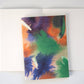 Das Papaya Paper Products | Notizheft A6 mit Aquarell/Watercolor-Motiv ruht mit seinem lebendigen, abstrakten Einband auf leeren Seiten eines aufgeschlagenen weißen Buches, die beide auf einer hellen Fläche angeordnet sind.