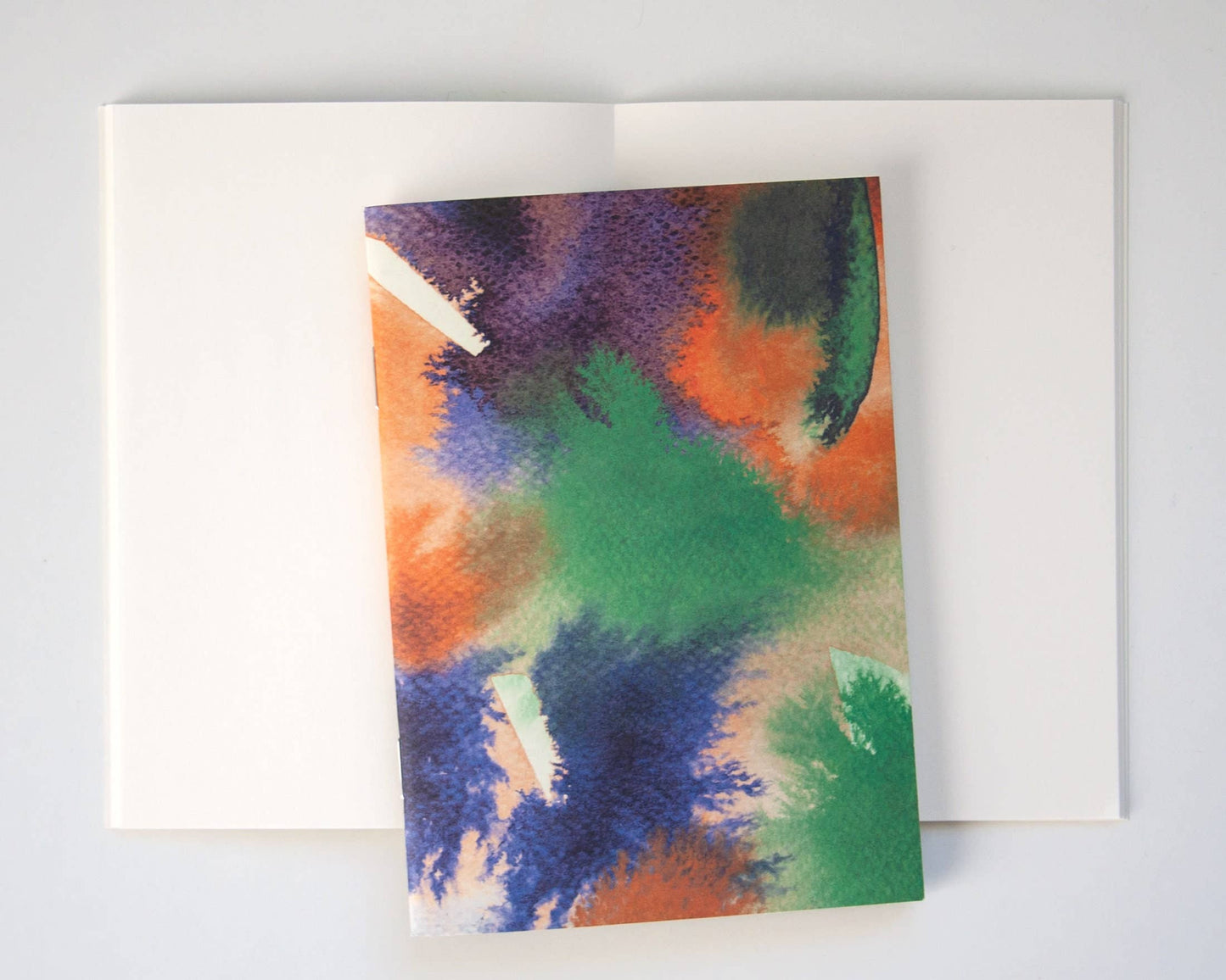 Das Papaya Paper Products | Notizheft A6 mit Aquarell/Watercolor-Motiv ruht mit seinem lebendigen, abstrakten Einband auf leeren Seiten eines aufgeschlagenen weißen Buches, die beide auf einer hellen Fläche angeordnet sind.