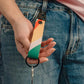 Eine Hand hält den Notabag-Schlüsselanhänger „Flow“ – einen farbenfrohen Schlüsselanhänger in Orange, Pfirsich, Weiß und Grün von Notabag –, der an der Gürtelschlaufe einer blauen Jeans befestigt ist und an dem Schlüssel an einem schwarzen Ring hängen.